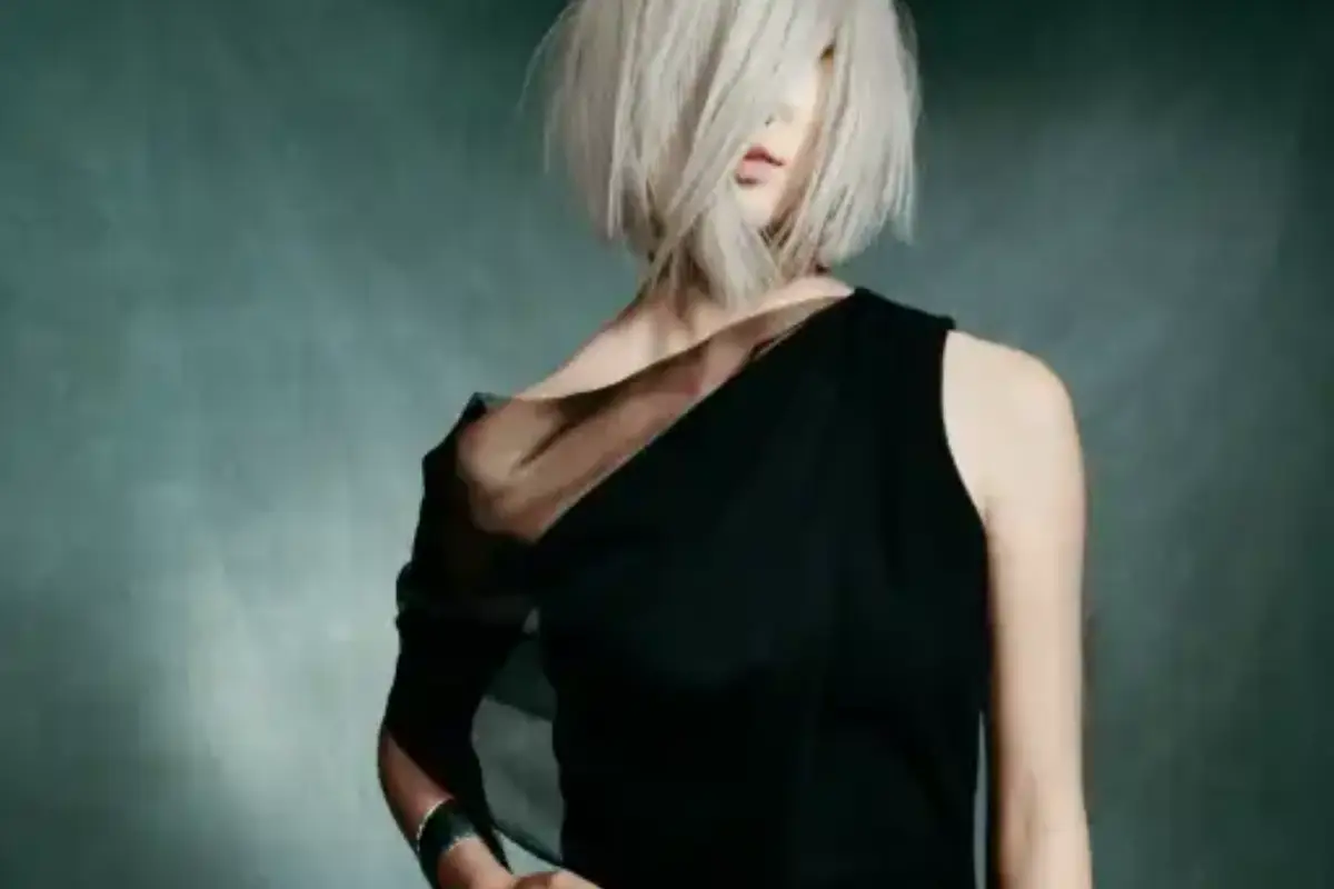 Person with chin-length platinum blonde hair wears a one-shoulder black dress and poses against a dark, blurred background. -Pure Salon, Ankeny, Johnston, & Indianola, IA