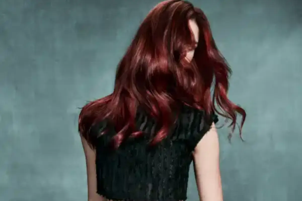 Person with long, wavy, dark red hair facing away from the camera, wearing a short-sleeved black top, against a gray background. -Pure Salon, Ankeny, Johnston, & Indianola, IA