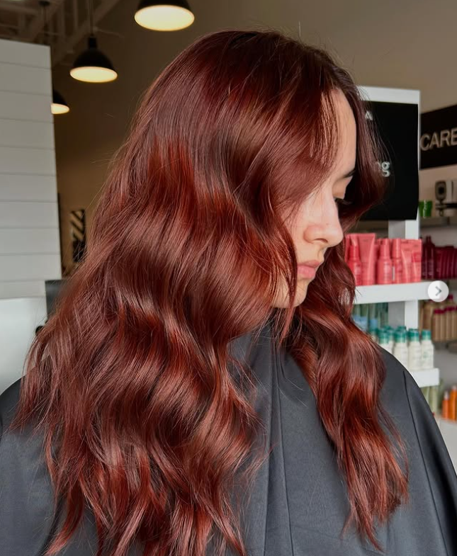 A person with long, wavy, auburn hair sits in a salon chair, wearing a black cape. Salon shelves with hair products are visible in the background. -Pure Salon, Ankeny, Johnston, & Indianola, IA