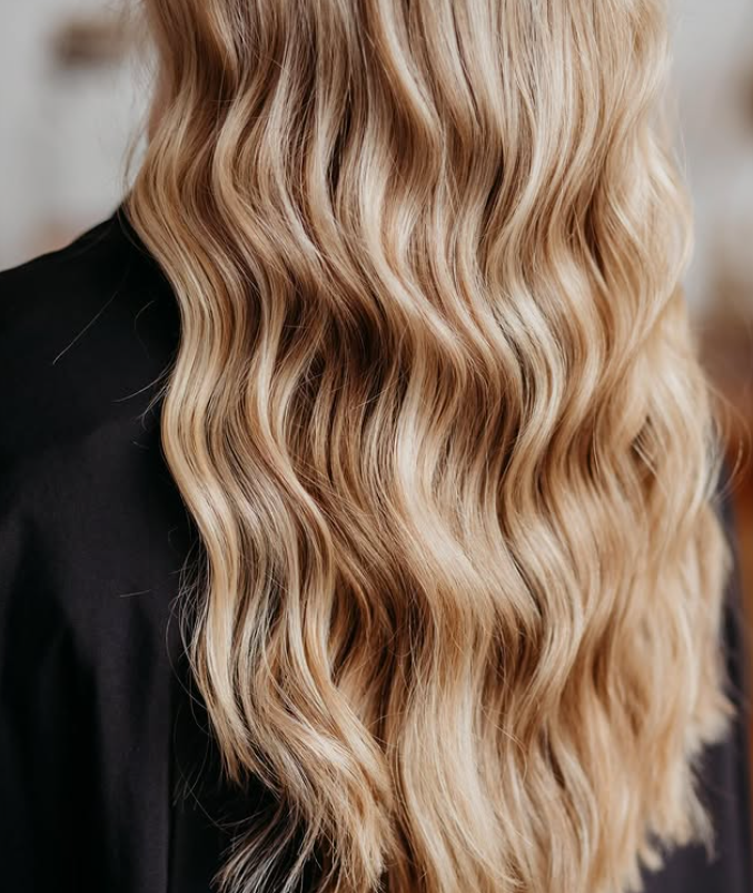 A person with long, wavy blonde hair wearing a black garment, shown from the back. -Pure Salon, Ankeny, Johnston, & Indianola, IA