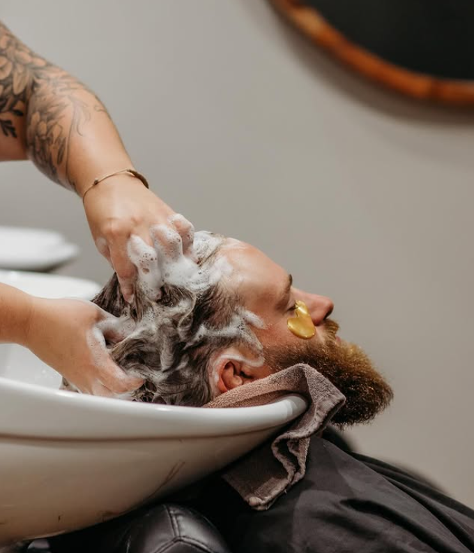 A person receives a hair wash with shampoo at a salon sink, while wearing under-eye gel patches. -Pure Salon, Ankeny, Johnston, & Indianola, IA