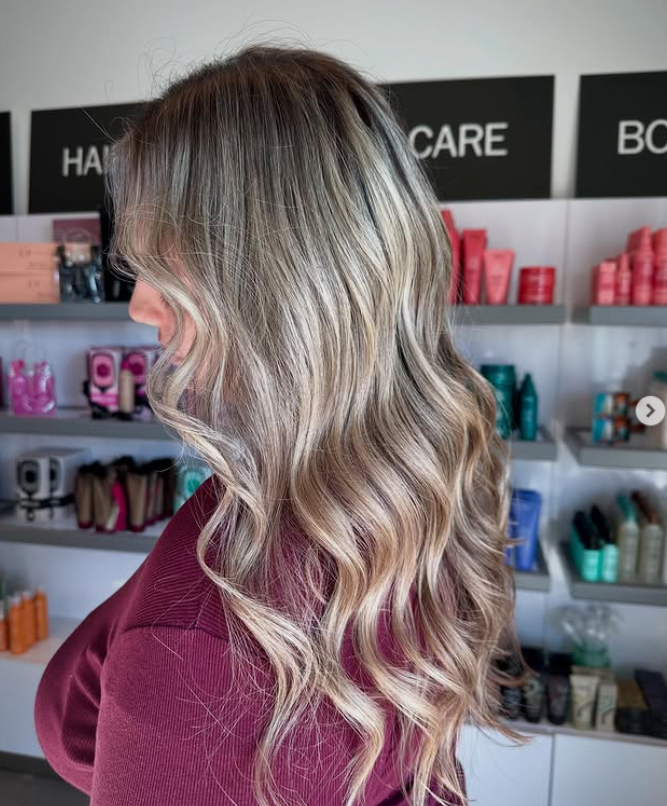A person with long, wavy, blonde-highlighted hair stands in a salon, facing sideways. Haircare products are displayed on shelves in the background. -Pure Salon, Ankeny, Johnston, & Indianola, IA