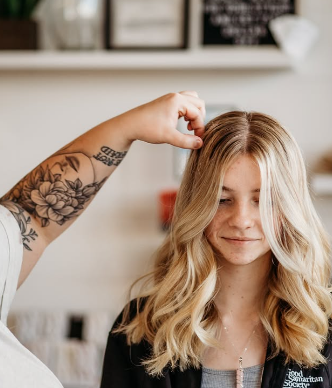 A hairstylist with a floral tattoo adjusts a young woman's wavy, blonde hair in a salon. -Pure Salon, Ankeny, Johnston, & Indianola, IA