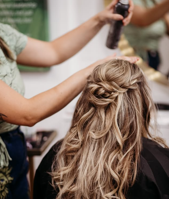 A person styles another person's wavy, half-up hair with hairspray in a salon setting. -Pure Salon, Ankeny, Johnston, & Indianola, IA