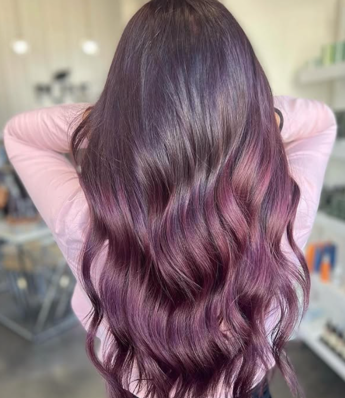 Person with long, wavy hair colored in shades of purple, wearing a light pink top, standing indoors with shelves and products blurred in the background. -Pure Salon, Ankeny, Johnston, & Indianola, IA