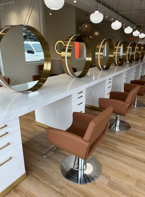 A row of brown salon chairs faces round gold-framed mirrors on a white counter in a modern, well-lit hair salon with wooden floors. -Pure Salon, Ankeny, Johnston, & Indianola, IA