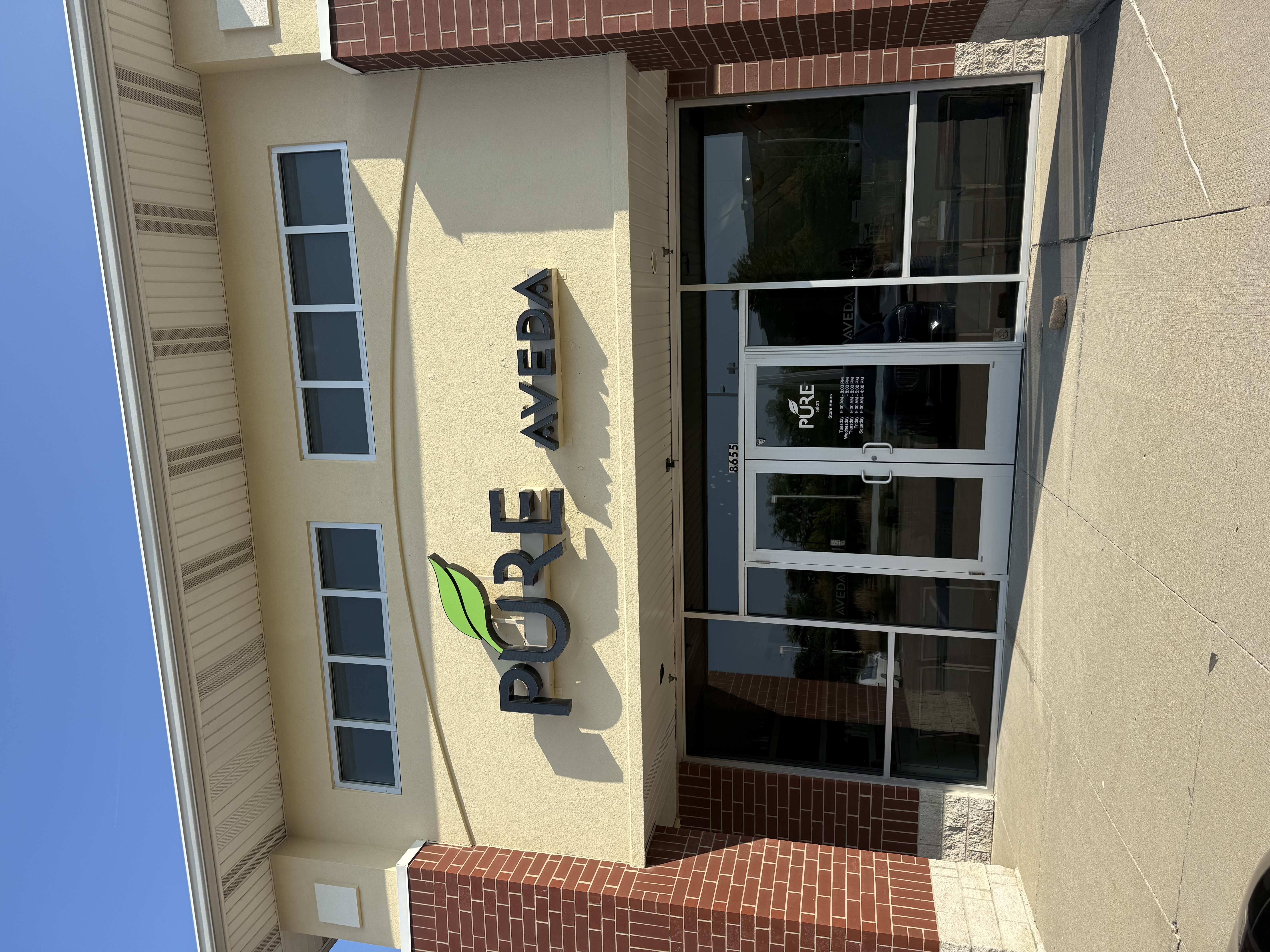 Storefront of Pure Aveda with glass doors and windows, beige and brick exterior, and sunlight casting shadows on the building. -Pure Salon, Ankeny, Johnston, & Indianola, IA