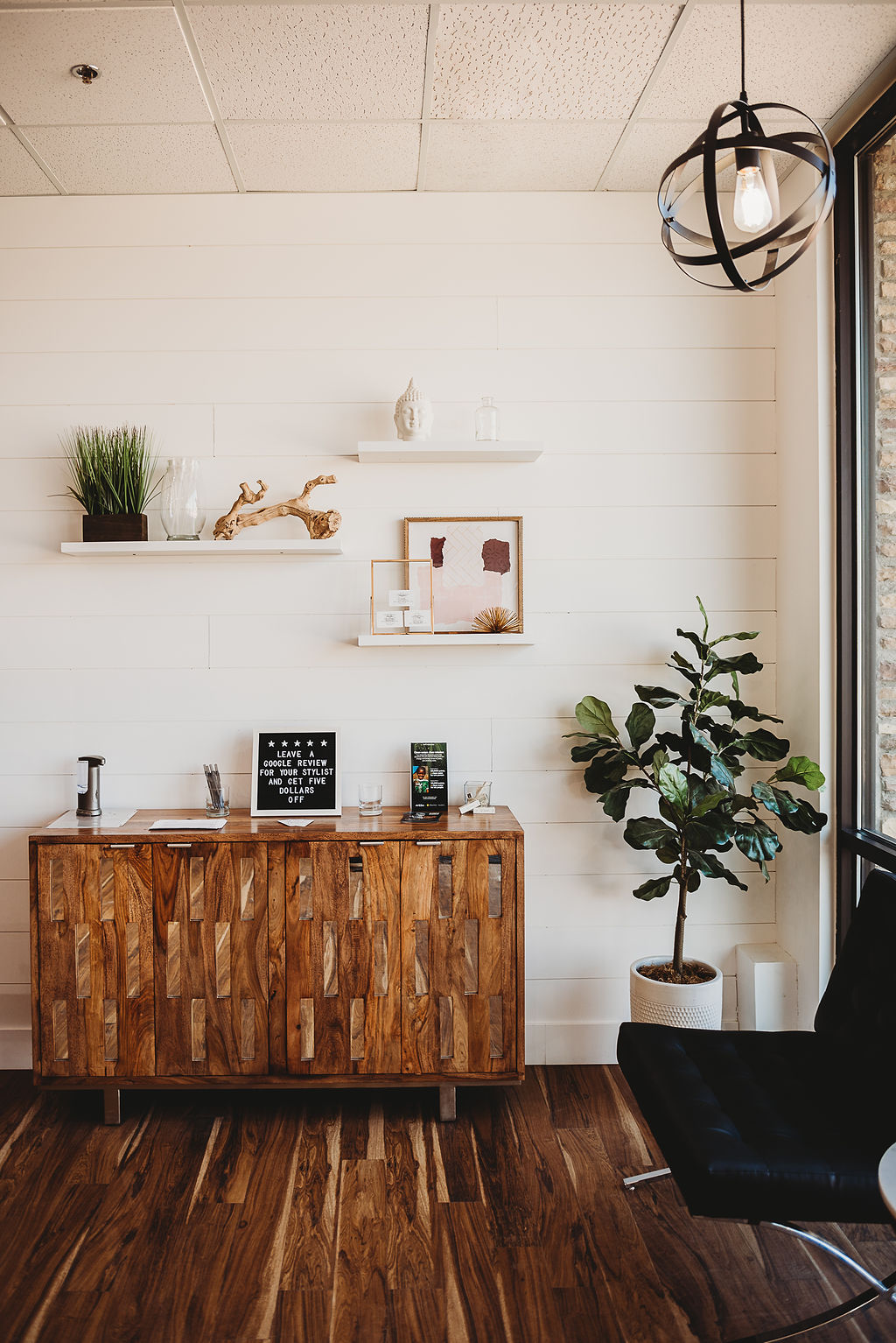 A modern office waiting area with a wooden sideboard, decorative shelves, a potted plant, and a black chair by a large window. -Pure Salon, Ankeny, Johnston, & Indianola, IA