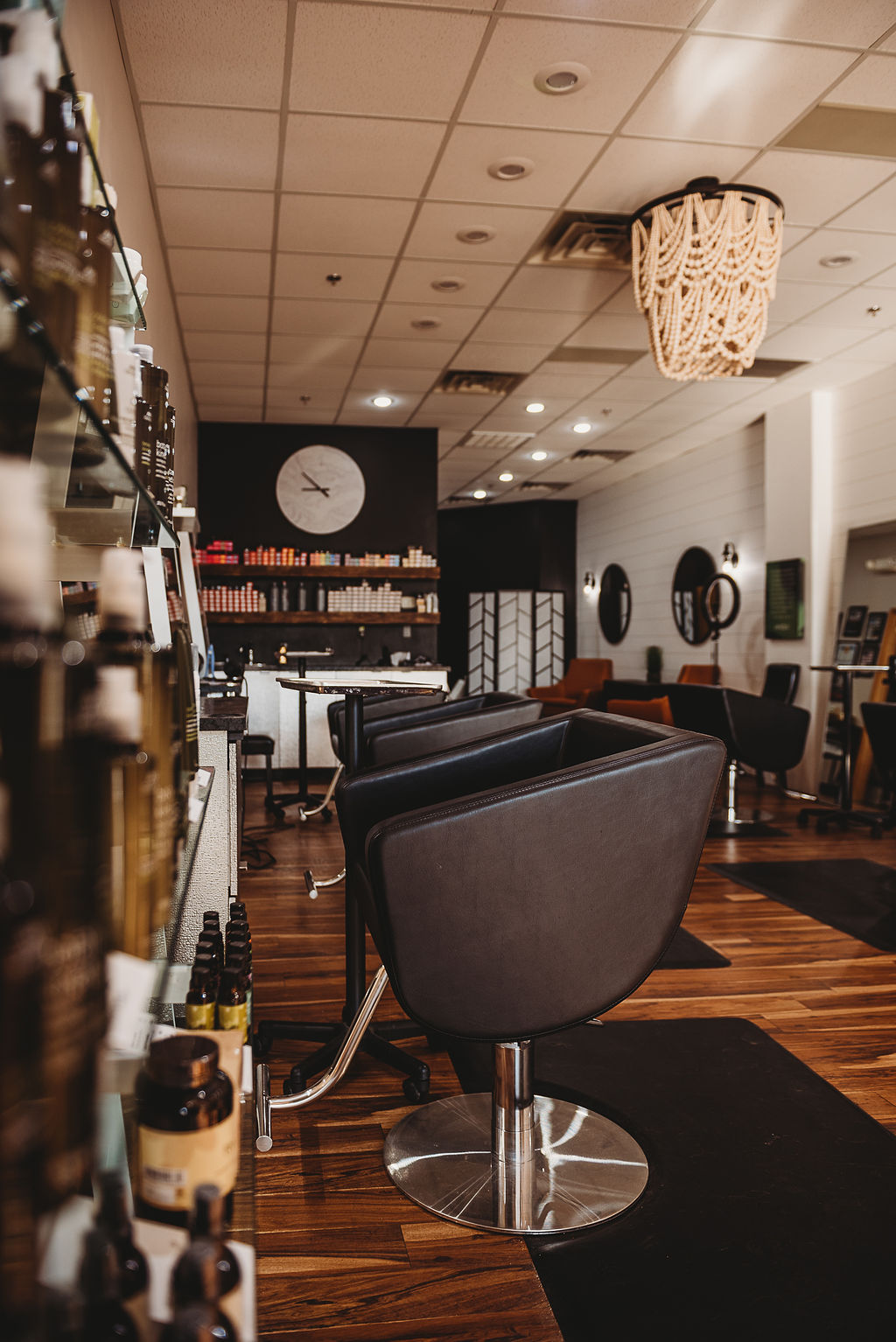 A modern hair salon interior with black chairs, wooden floors, shelves of hair products, round mirrors, and a decorative chandelier. -Pure Salon, Ankeny, Johnston, & Indianola, IA