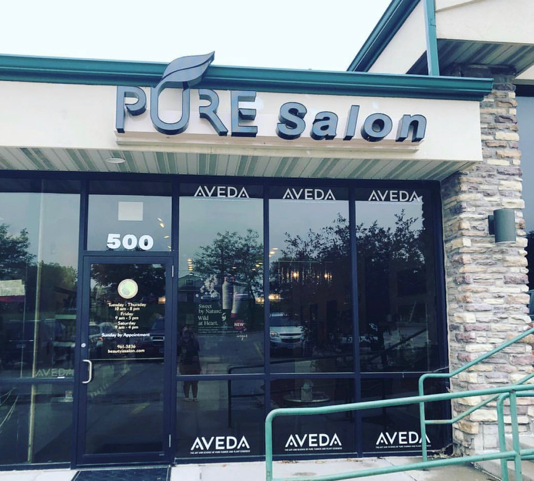 Storefront of PURE Salon with large windows, AVEDA branding, a glass door with business hours, and a stone accent wall on the right. -Pure Salon, Ankeny, Johnston, & Indianola, IA