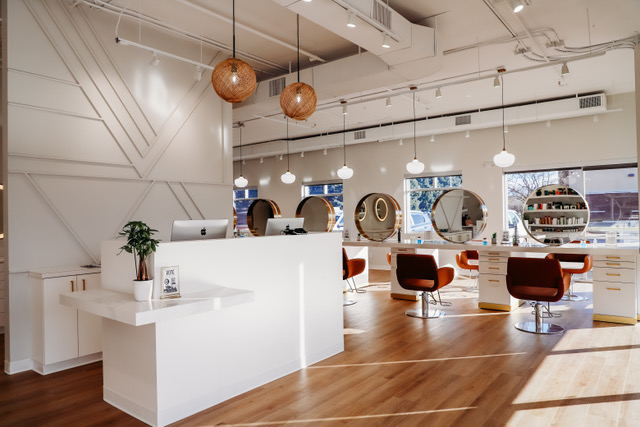 Modern hair salon interior with white counters, circular mirrors, brown chairs, pendant lights, wooden flooring, and large windows allowing natural light. -Pure Salon, Ankeny, Johnston, & Indianola, IA